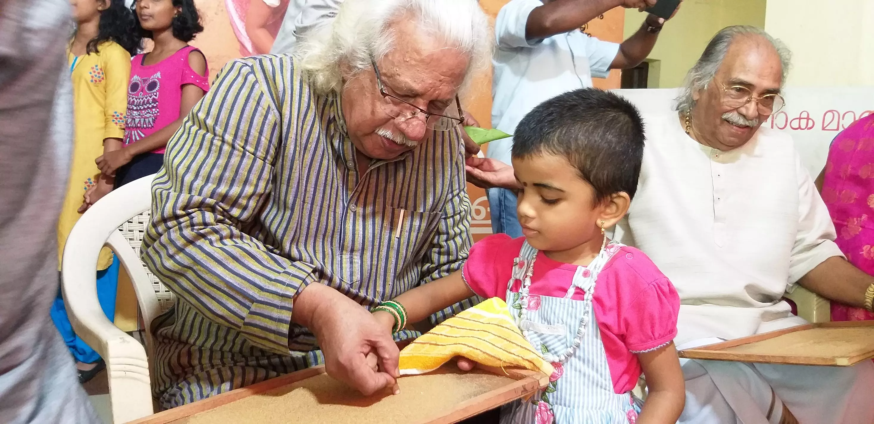 Adoor Gopalakrishnan helps a little one write Malayalam alphabets on a sand-tray.