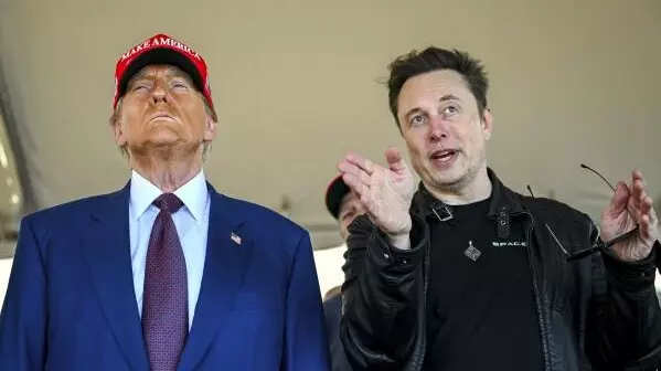 A picture of Donald Trump on the left with a blue suit and tie on with a red MAGA cap and to the right is Elon Musk in a black leather jacket and black t-shirt, Musk is gesturing his palm outward like he is pointing out something to Trump.