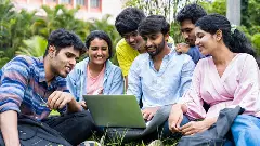 A group of students sit on a lawn and look into a laptop with expectant smiles.