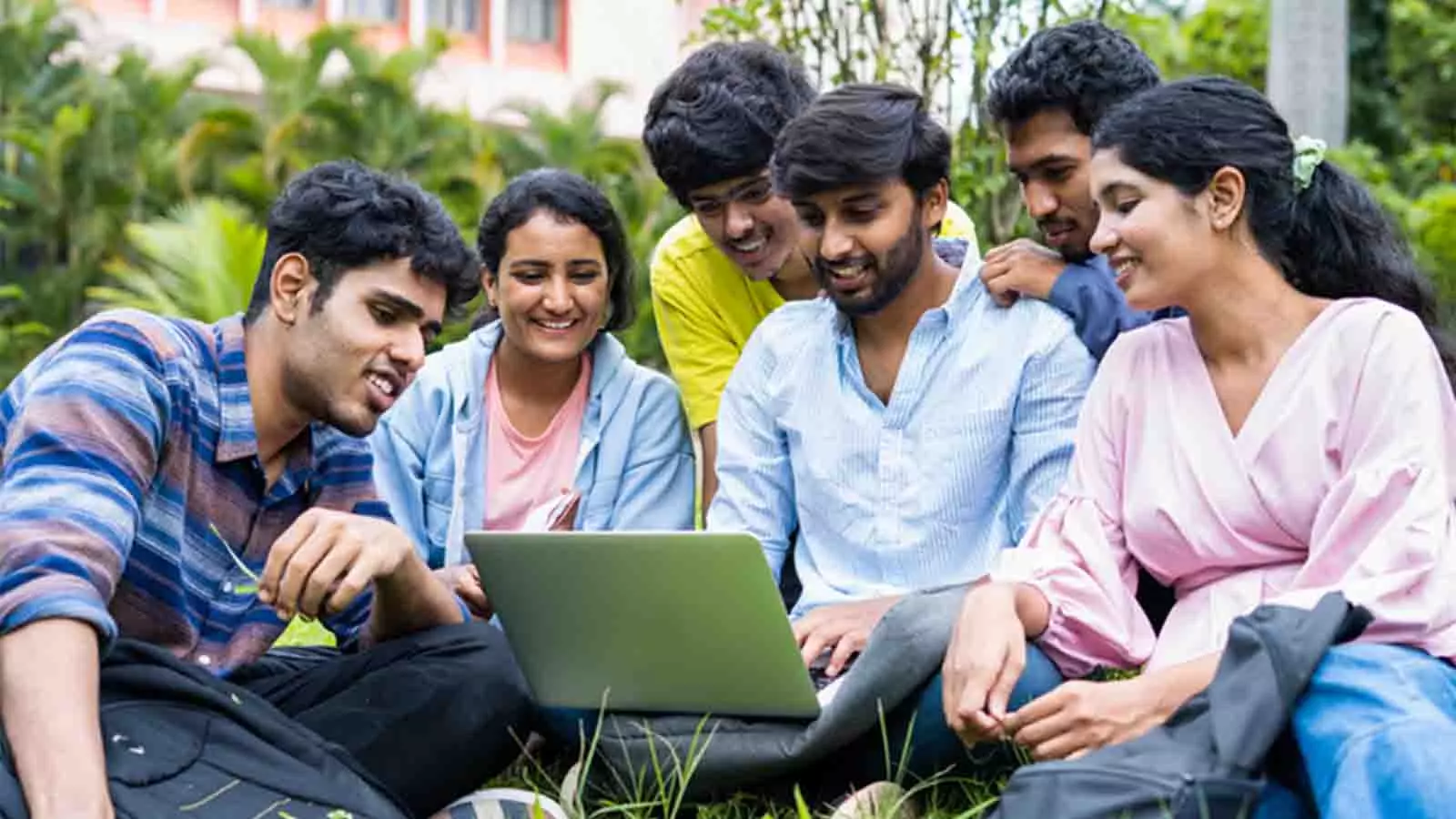 A group of students sit on a lawn and look into a laptop with expectant smiles.