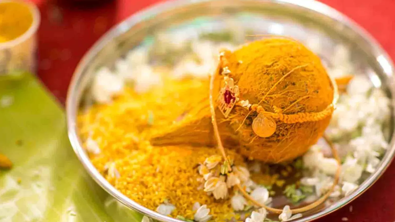 A coconut with turmeric on top has a wedding thali or mangalsutra tied to it and placed on a silver plate next to yellow rice grains and jasmine flowers on a red tablecloth with a banana leaf to the side