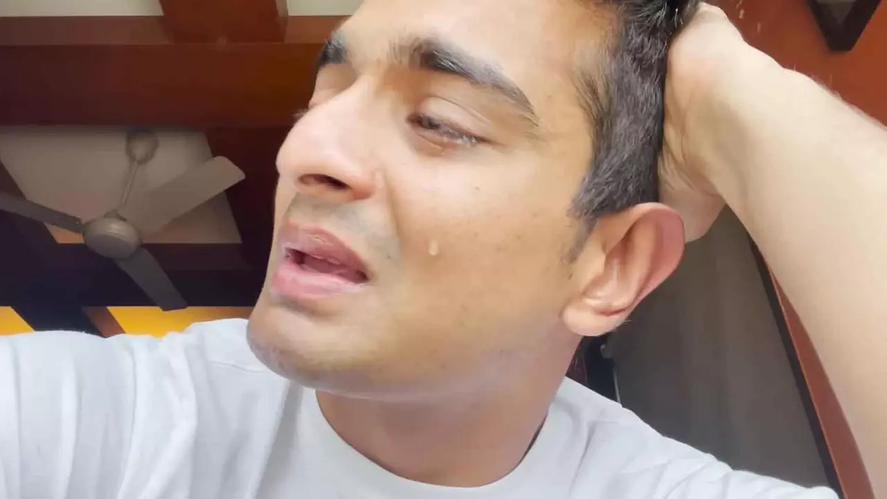 A video screengrab of Ranveer Allahbadia with his face in profile, a single tear falling down his cheek as he sits in what looks like his bedroom with a white t-shirt on and his hand on his head looking distressed mid-talk.