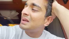 A video screengrab of Ranveer Allahbadia with his face in profile, a single tear falling down his cheek as he sits in what looks like his bedroom with a white t-shirt on and his hand on his head looking distressed mid-talk.